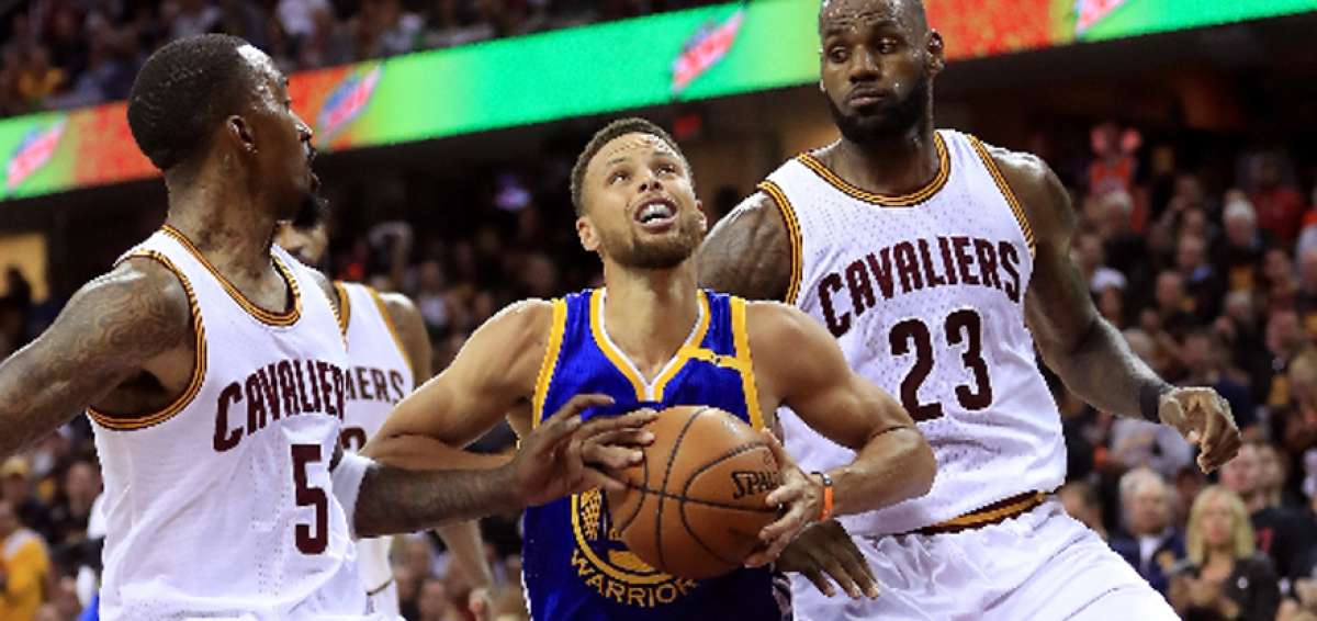 Stephen Curry (c) controla el bal&oacute;n y mira al tablero antes de realizar el lanzamiento, dos jugadores observan.