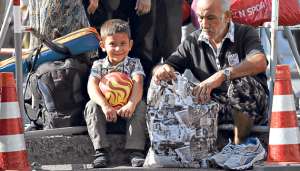 Un ni&ntilde;o sonr&iacute;e junto a su padre, quien revisa su equipaje en una de las islas griegas, luego de llegar procedente de Turqu&iacute;a.  El 60% de la poblaci&oacute;n que llega no recibe asistencia m&eacute;dica ni sanitaria. Foto: AFP
